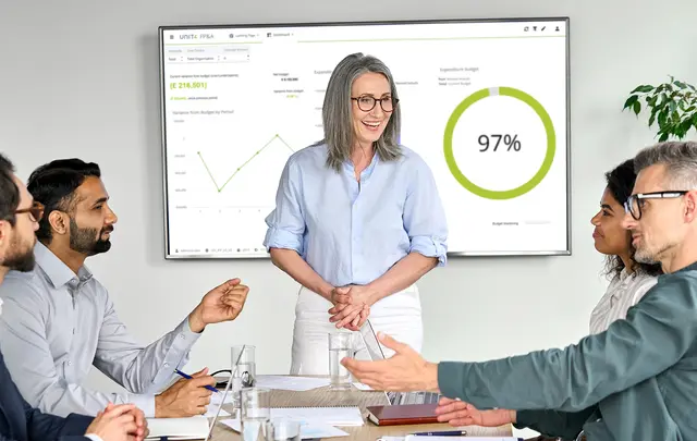Smiling woman talking to people around a table. Behind her a screen with Unit4 FPA