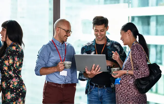 Four people gathered around a laptop, smiling and chatting in a modern office setting