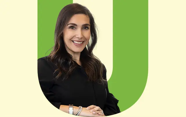 Michelle MacCarthy A smiling woman with long dark hair wearing a black shirt and bracelets, posing against a light green background. 