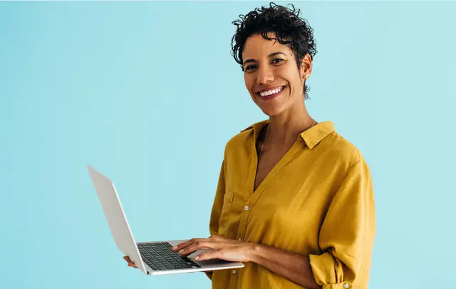 A woman with curly hair smiles while holding a laptop in front of a blue background