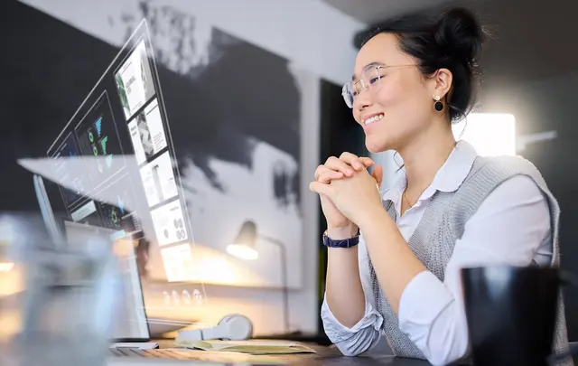 A woman smiling while sitting at a desk with a computer screen displaying charts and graphs. 