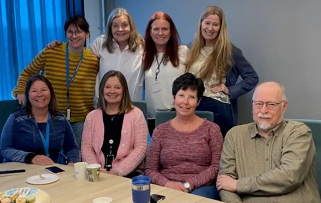A group of eight people smiling around a table with coffee cups and a cake. 