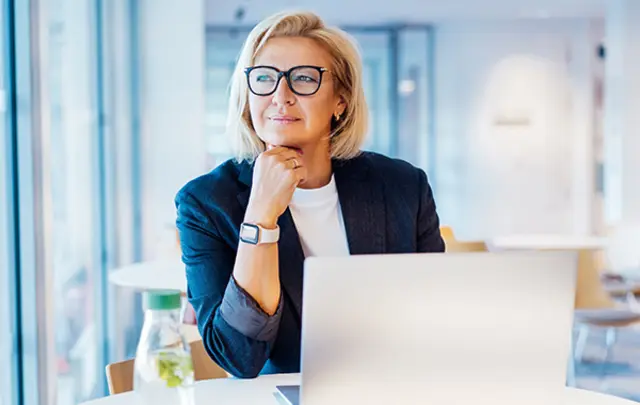A thoughtful woman with blonde hair and glasses sits by a laptop in a modern workspace. 