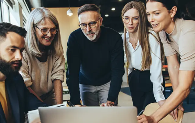 A group of five people gathered around a laptop, discussing ideas in an office setting