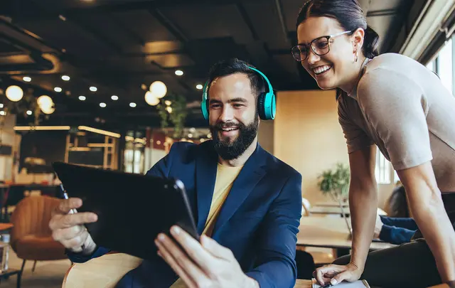 A man in a blue jacket and turquoise headphones smiles while using a tablet, with a woman beside him