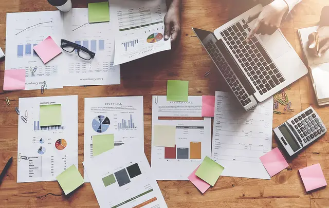 A wooden table with financial documents, a laptop, sticky notes, a calculator, and a notebook. 