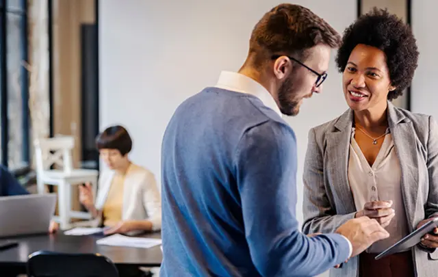 A man and woman discuss over a tablet in a bright office, with coworkers in the background