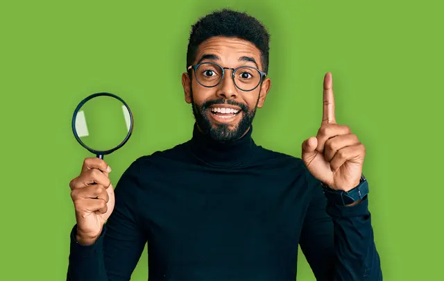A smiling man in a black turtleneck holds a magnifying glass in one hand and points up with the other against a green background. 