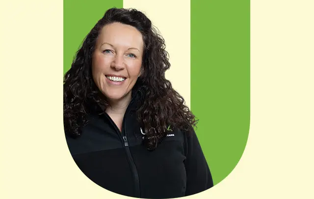 A woman with curly hair wearing a black zip-up jacket, smiling against a light green background. 