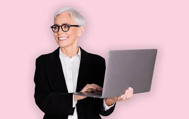 A smiling older woman with glasses stands holding a laptop in front of a pink background. 