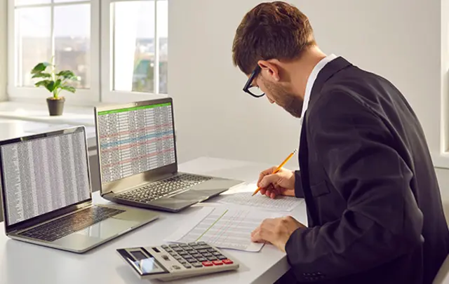 A man in a suit working at a desk with two laptops, a calculator, and paperwork. 