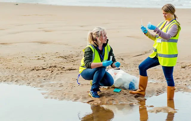Two women in yellow vests collecting samples and cleaning the beach, with trash bags nearby.