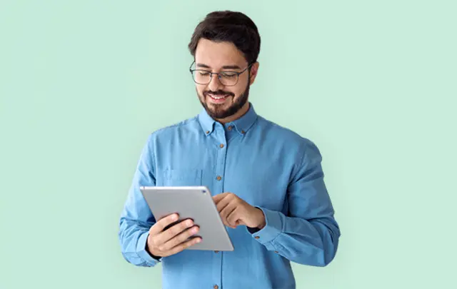 A man in a blue shirt smiles while using a tablet in front of a light green background. 