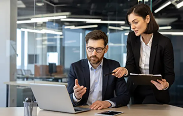 A man in glasses sits at a laptop while a woman in business attire stands beside him, pointing and discussing. 