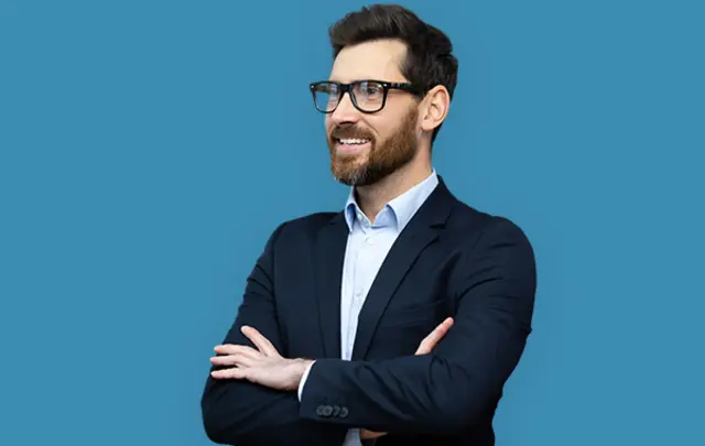A man in a dark suit and glasses smiling, with arms crossed in front of a blue background. 