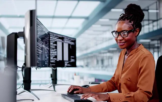 A professional seated at a desk in a modern office, working on a dual-monitor setup displaying code or text, wearing glasses and a brown shirt, smiling and engaged in their task.