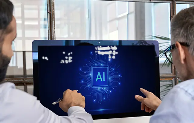 Two individuals collaborating in an office, focusing on a computer screen displaying a digital circuit board with 'AI' prominently featured, symbolizing artificial intelligence and teamwork.