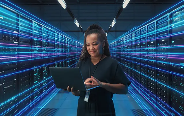 A woman in a black outfit using a laptop in a server room with glowing blue lights. 