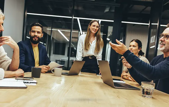 A group of five people in a meeting room, smiling and discussing at a table with laptops and drinks. 