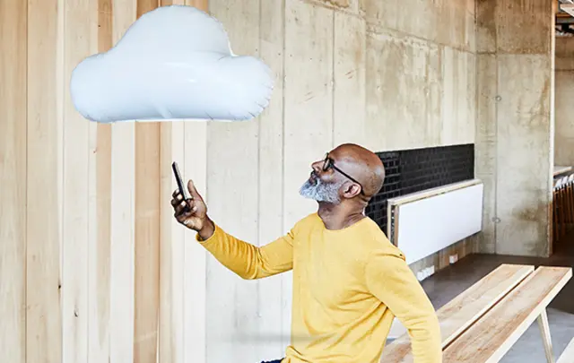 A man in a bright yellow sweater and glasses holds a smartphone while looking at a white cloud-shaped balloon in a modern indoor space with wooden and concrete elements.