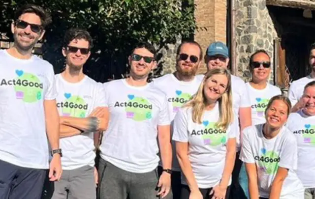 Group of nine people outdoors, wearing matching white t-shirts with 'Act4Good' printed on them, posing together in two rows.