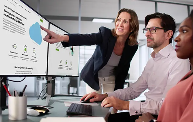 A professional office setting with three colleagues collaborating. One person stands, pointing at a monitor, while two others are seated, focused on the screens