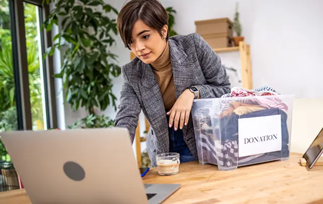A person in a gray blazer leans over a wooden table, focused on a laptop.