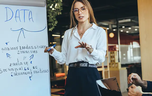 A professional setting featuring a person in a white shirt and glasses presenting data on a whiteboard, with 'DATA' written prominently at the top. 