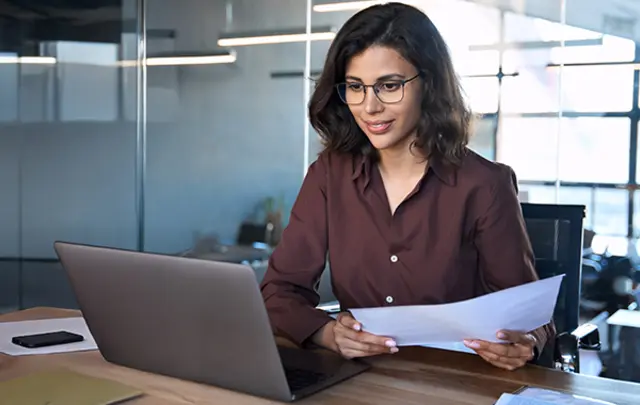 A woman wearing glasses and a brown shirt sits at a wooden desk in a modern office.