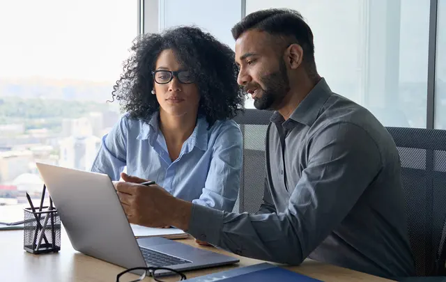 Two professionals seated at a desk in a modern office, focused on a laptop. 