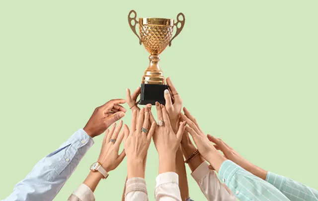 A gold trophy with decorative handles and a black base is centered in the image, surrounded by multiple hands of diverse skin tones and clothing styles reaching upward. 