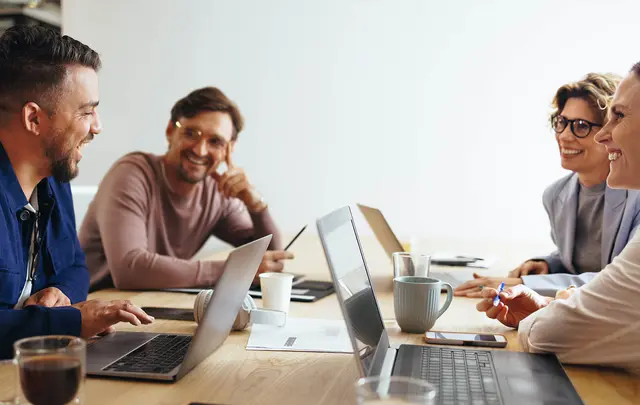 Five individuals gathered around a light wooden table in a modern office setting, engaged in a collaborative discussion. 