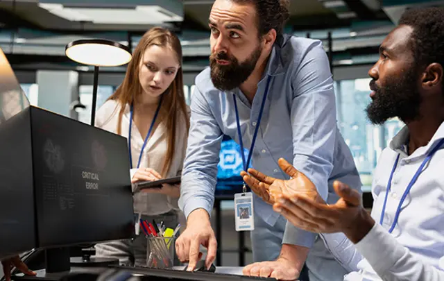 Three professionals in a modern office collaborate around a computer monitor displaying a 'CRITICAL ERROR' message. 