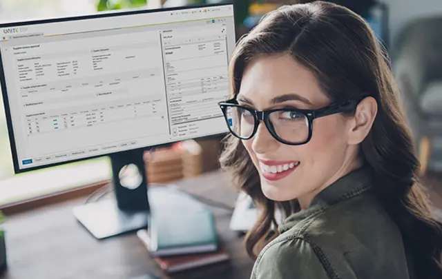 A smiling woman with black-framed glasses sits at a wooden desk in a modern, well-lit office. 