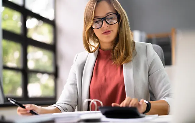 A professional woman with light brown hair and black-framed glasses works at a desk in a bright office.