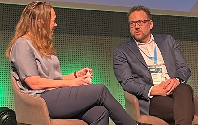 Two professionals seated in modern armchairs on a stage, engaged in a panel discussion.