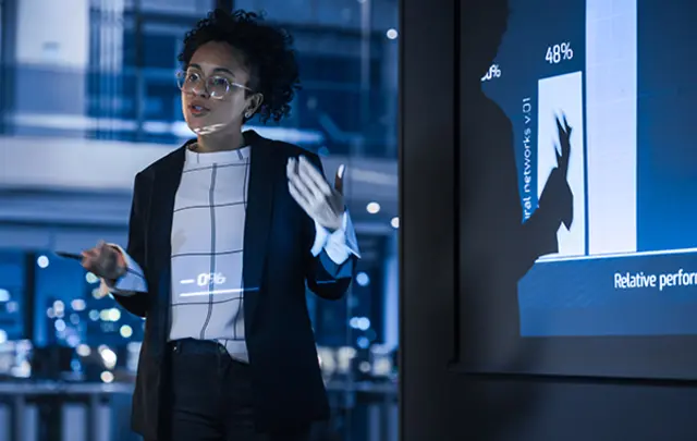 A professional presentation scene featuring a woman with dark curly hair and glasses, gesturing while explaining a bar chart projected on a screen.
