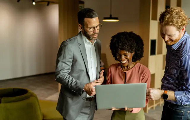 Three colleagues gathered around a silver laptop in a modern office setting.