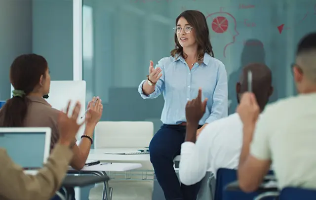 A professional and educational setting with a woman seated on a desk at the center, gesturing as she leads a discussion