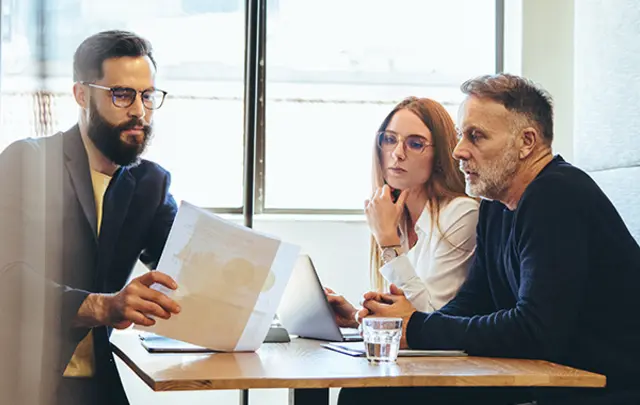 Three individuals seated around a wooden table in a professional setting, engaged in a collaborative discussion.
