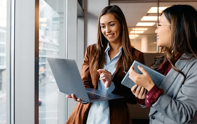 Two professionals in business attire stand near a large window with an urban cityscape in the background.