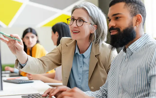 A professional setting with four individuals, where a woman with gray hair and glasses points at a computer screen with a pen, smiling. 