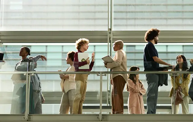 A diverse group of professionals interact on a modern, glass-railed walkway in a bright, naturally lit building. 