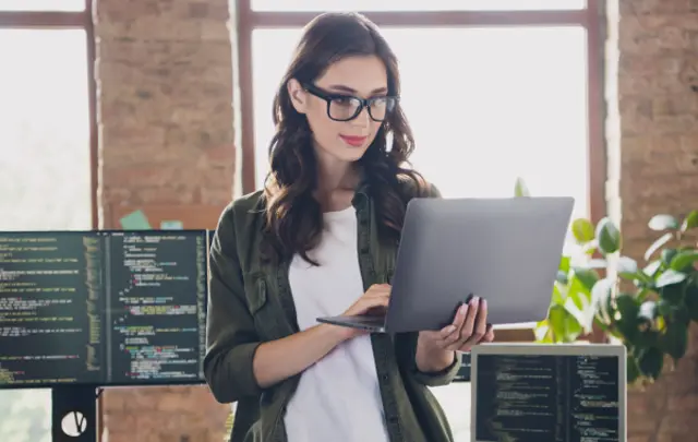 A young woman with long dark brown hair and glasses works intently on a silver laptop in a modern office.