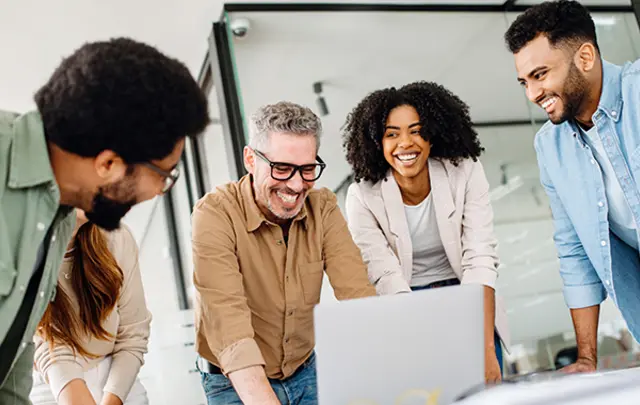 Five professionals gather around a silver laptop on a white table in a modern, well-lit office.