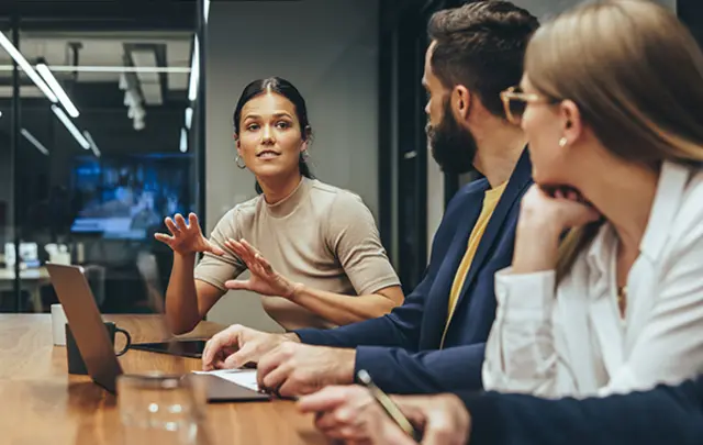 Three colleagues engaged in a business discussion around a polished wooden table in a modern office.