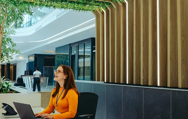 A smiling woman with reddish-brown hair and glasses sits at a white desk in a modern office lobby. 