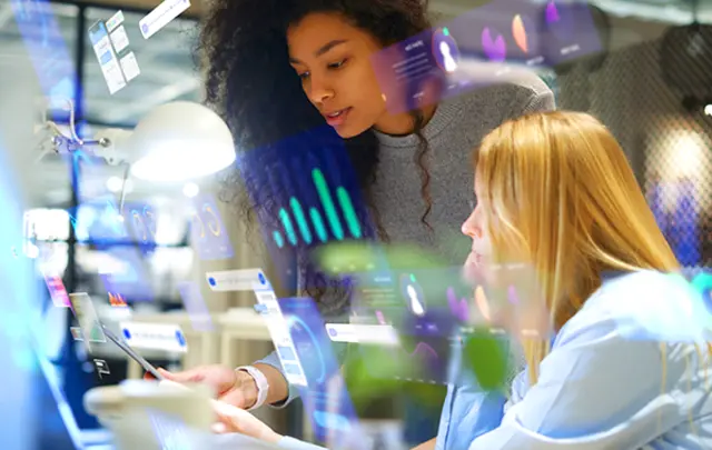 Two women collaborate in a modern office, surrounded by glowing holographic data visualizations.