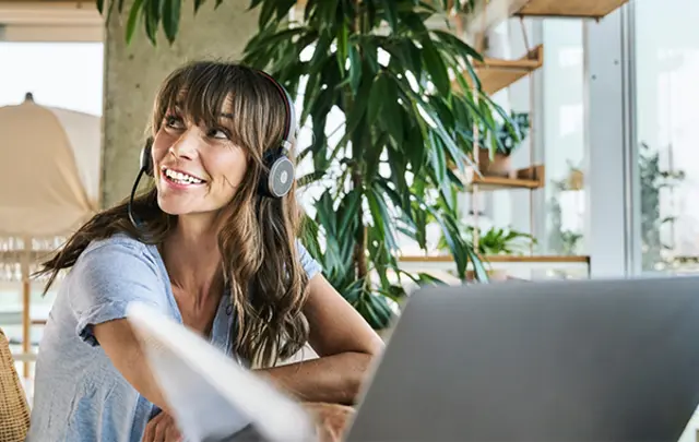 A smiling woman with wavy brown hair and headphones with a microphone sits at a desk with a silver laptop. 