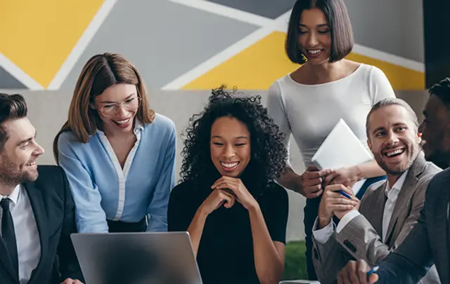 Six diverse professionals collaborate around a laptop in a modern office. 